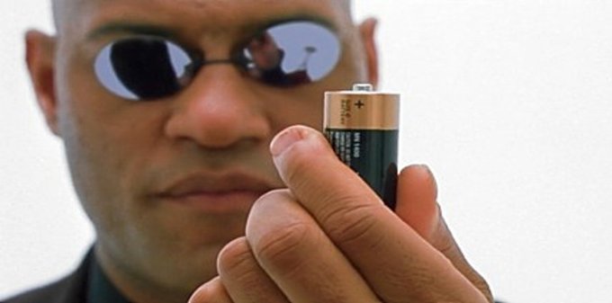 A close-up of a man wearing reflective sunglasses holding a small battery in front of his face, reminiscent of Morpheus from The Matrix.