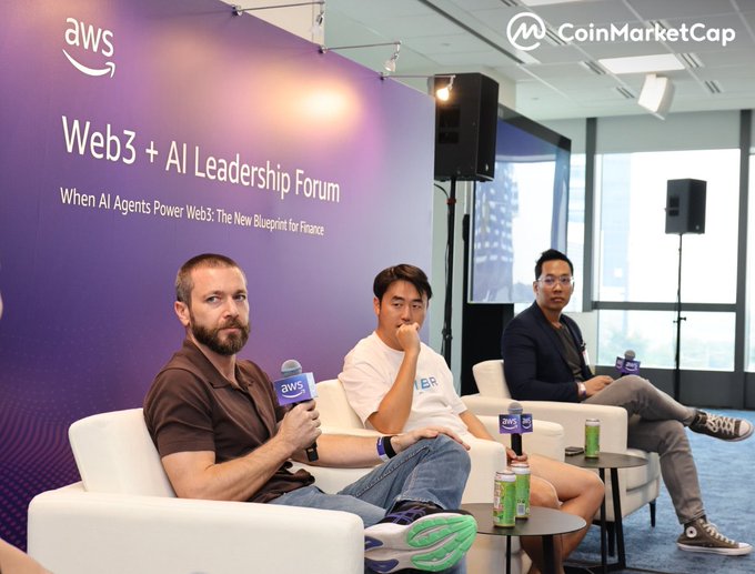 Three men sit on a stage at the "Web3 + AI Leadership Forum" hosted by AWS and CoinMarketCap, with the subtitle "When AI Agents Power Web3: The New Blueprint for Finance," engaged in a panel discussion with AWS-branded microphones. The image captures the live setting referenced in the post about CoinMarketCap's CEO joining AWS, Amber Group, and OpenAI to discuss moving AI × crypto from demo to production at TOKEN2049.