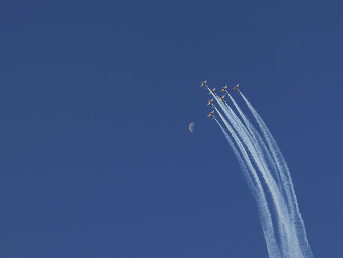 A formation of six fighter jets flies through a deep blue sky leaving white contrails behind them, with a crescent moon visible in the background.