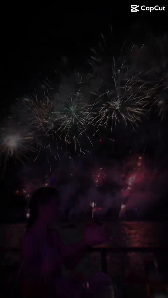 The image shows a woman standing next to a railing with her arms outstretched towards the fireworks display happening above them. The fireworks are exploding in the night sky, creating a beautiful spectacle of colors and patterns. The scene captures the excitement and wonder of the moment as the fireworks light up the dark sky.