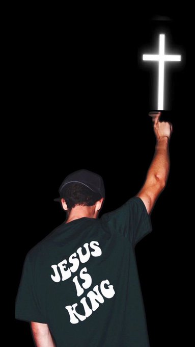 A person viewed from behind wearing a green shirt with "JESUS IS KING" printed on it, raising a glowing cross against a black background.
