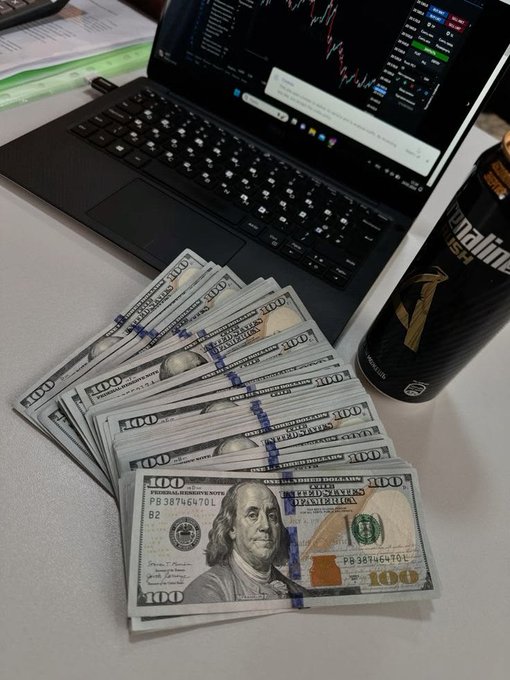 A fanned-out stack of $100 bills rests next to an energy drink and a laptop displaying stock charts, visually representing the milestone of hitting the first $100K in trading or investing.