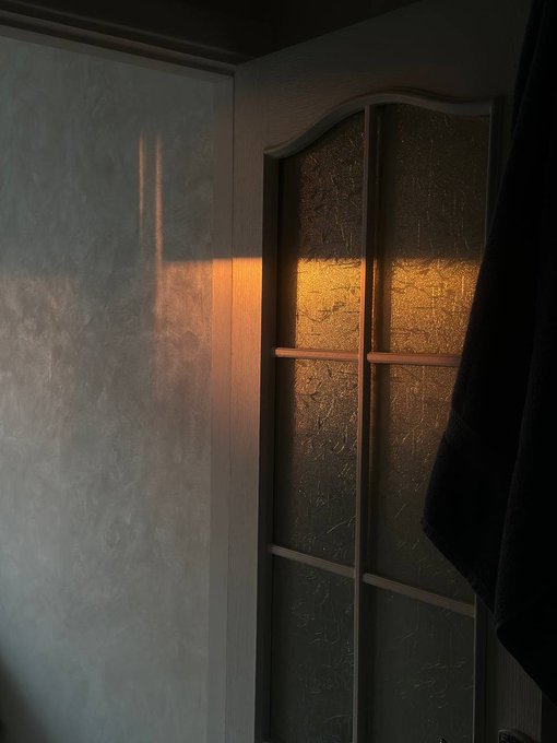 Sunlight streaming through a textured glass window pane, casting a warm golden glow onto a nearby wall, with a dark curtain partially visible on the right.