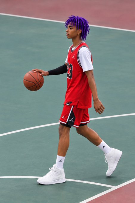 A young basketball player with purple dreadlocks, wearing a red and white Jordan jersey with the number 3, dribbles a basketball on an outdoor court.