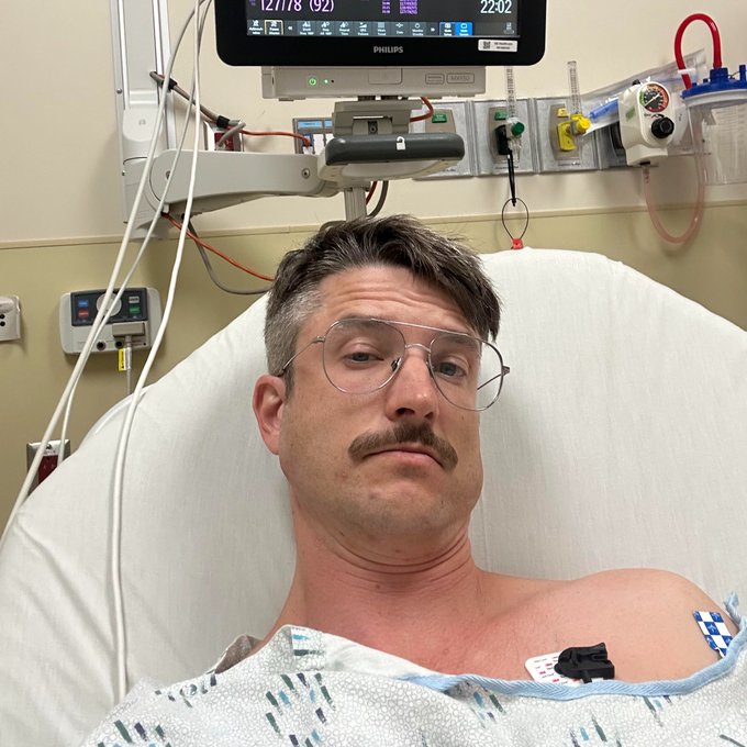 A man with a mustache and glasses lying in a hospital bed with medical monitors and equipment visible behind him.