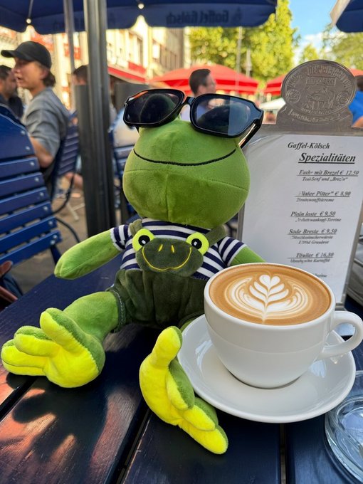 A plush frog wearing sunglasses sits at an outdoor café table next to a latte with latte art, while a menu advertises "Gaffel-Kölsch Spezialitäten" including drinks like "Köbes Pitter," "Pilsa Josta," and "Süße Brezze" priced from €5.50 to €12.50—fitting the "$FROG" reference as a playful frog-themed café moment.