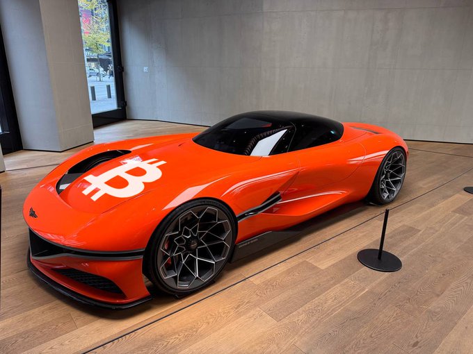 An orange sports car with a Bitcoin logo on the hood is displayed in a modern showroom with wooden flooring and concrete walls.