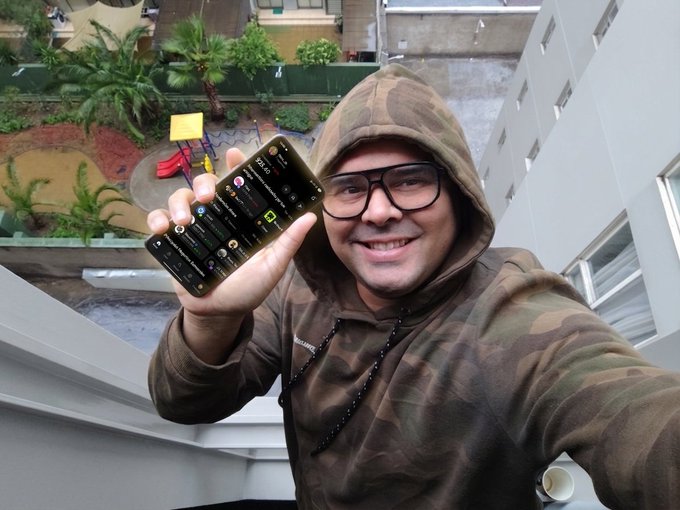 A man in a camouflage hoodie and glasses takes a selfie on a balcony, holding up a smartphone showing a messaging app.