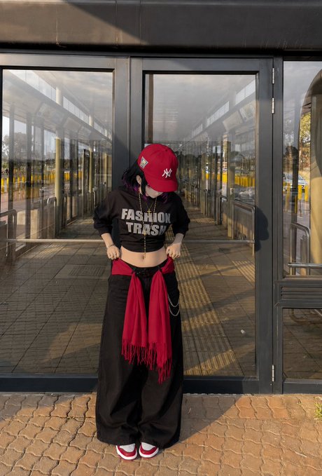A person stands outside glass doors wearing a red NY Yankees cap, a black 'FASHION TRASH' cropped hoodie, and baggy black pants with a red scarf tied at the waist.