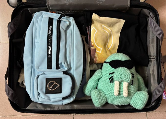 An open suitcase contains a light blue sling bag with 'Ready Set Play' text, a green crocheted plush toy with tusks, and a yellow shirt featuring a white logo.