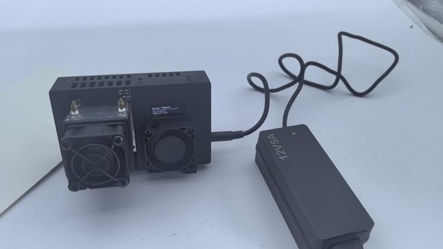 A black electronic device with two cooling fans and an attached power adapter labeled 'AGL' sits on a white surface.