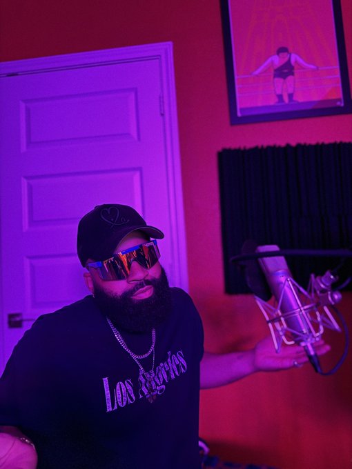 Bearded man wearing sunglasses, a dark 'Los Angeles' t-shirt, and a cap sitting in a studio lit with pink/purple lighting, next to a microphone, with a framed wrestling photo on the wall behind him.