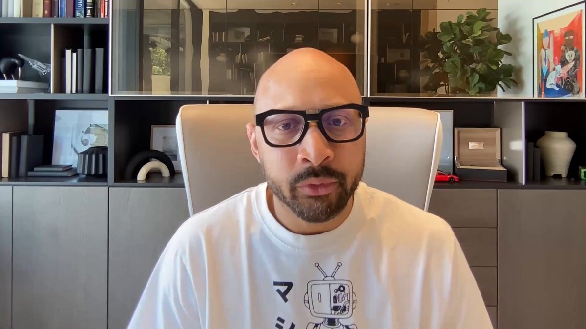 The image shows a man wearing glasses sitting at a desk in front of a bookshelf filled with various items, including several books and vases. The man appears to be looking directly into the camera while wearing his glasses. There are also two potted plants visible in the scene - one on the left side of the bookshelf and another on the right side.