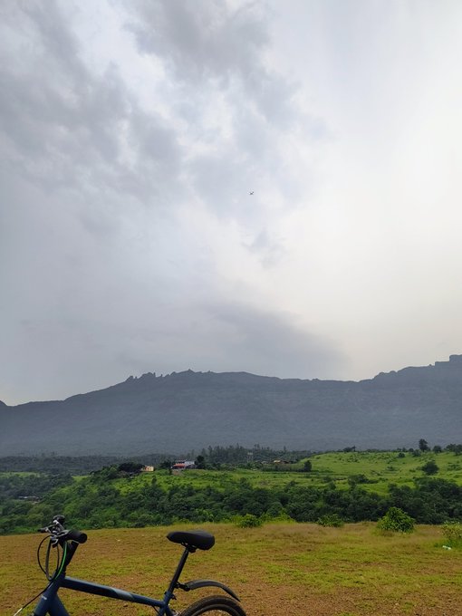 A scenic landscape photo featuring a cloudy sky, a mountain range, lush green hills with small buildings, and a bicycle parked in the foreground on a dirt area.