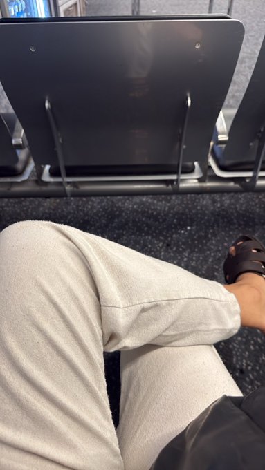 A first-person perspective photo taken while sitting in what appears to be an airport or bus terminal, showing the person's legs in beige pants and black sandals, with empty black chairs visible ahead.