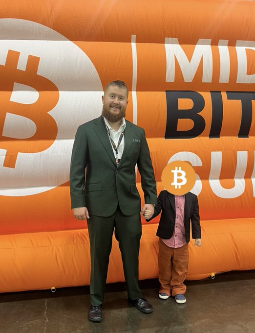 A man in a green suit stands next to a small figure with a Bitcoin logo as its face, posing in front of an orange Bitcoin-themed backdrop reading 'MID BIT...'