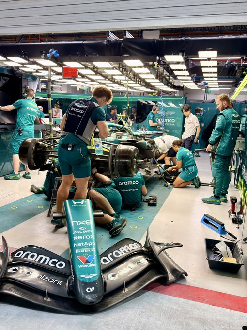 An Aston Martin F1 pit crew in teal Aramco uniforms services a race car in the garage, with visible sponsor logos including Aramco, NCE, Coinbase, Xerox, NetApp, Pirelli, and Valvoline displayed on the car's nose cone. The scene captures the behind-the-scenes VIP pit experience mentioned in the post, where guests watch Aston Martin's race operations up close while enjoying the hospitality.
