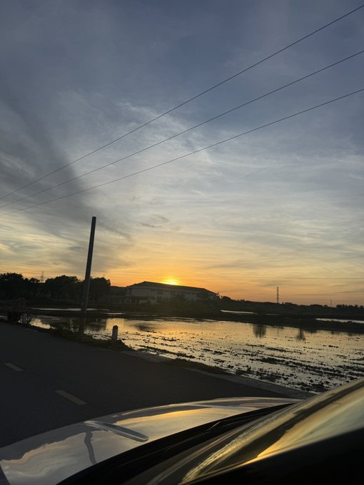A scenic sunset photo taken from inside a car, showing the sun setting over rice paddies and distant hills under a cloudy sky with power lines overhead.