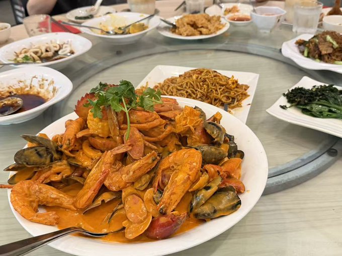 A large white plate piled high with shrimp, mussels, and vegetables in a spicy orange sauce sits on a round dining table surrounded by other dishes of food.