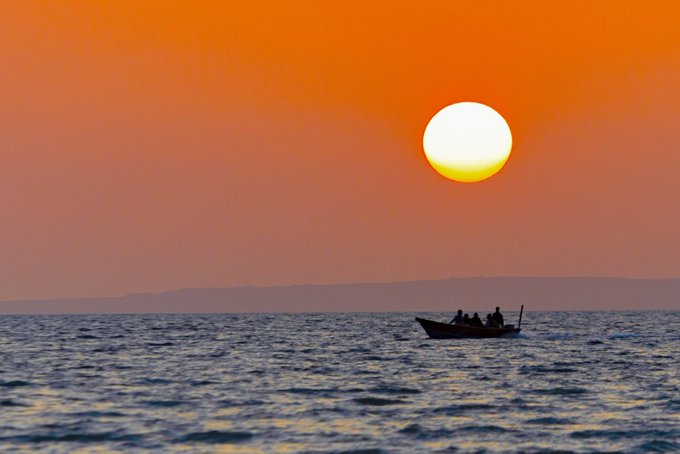 A serene photograph of a small boat with silhouetted figures on calm water under a glowing orange sunset with the sun low on the horizon.