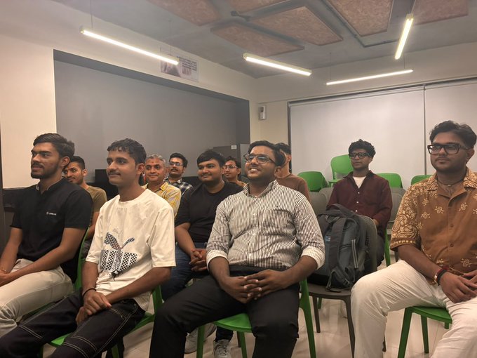 A group of young men sitting on green chairs in a meeting room or classroom, attentively watching something off-camera.