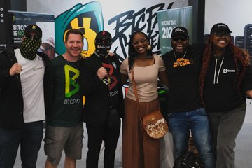 The image shows a group of six people standing together at a convention or event, posing for a photo. They are all wearing black clothing, which includes hats and shirts with Bitcoin written on them. The backdrop features a large sign that says "BTCPay", indicating that they are promoting Bitcoin as the theme of the event. One person in the group is holding up a sign that reads "BETTA".
