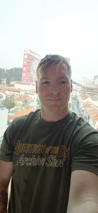 A man wearing a green t-shirt reading 'QUESTION OF THE DAY Archive Show' takes a selfie overlooking a coastal town with buildings.