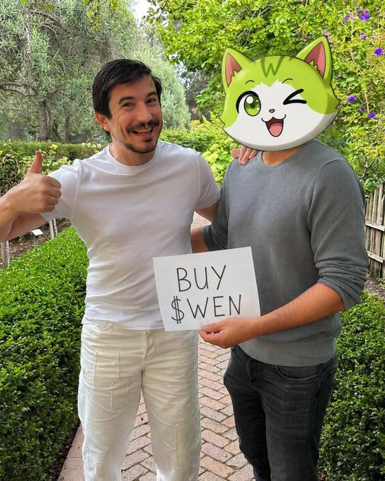 A man in a white t-shirt giving a thumbs-up next to another person holding a sign that reads "BUY $WEN," with a cartoon green cat face covering their identity.