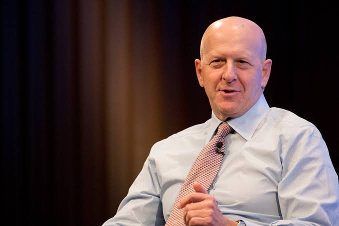 A bald man in a light blue dress shirt and patterned tie gestures while speaking, set against a dark curtained backdrop typical of a stage or interview setting. The image appears to depict a Goldman Sachs representative, connecting to the post about Goldman Sachs leading institutional holdings in U.S. spot XRP ETFs with $87.4M amid accelerating institutional crypto adoption.
