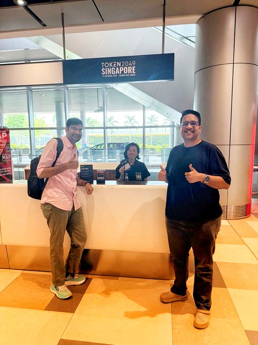 Two men pose smiling with thumbs up at a registration counter for the TOKEN2049 SINGAPORE cryptocurrency conference, with a staff member visible behind the desk.