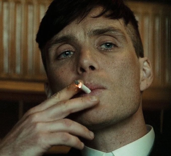 A close-up shot of a young man with dark hair smoking a cigarette, looking off to the side in a dimly lit room.