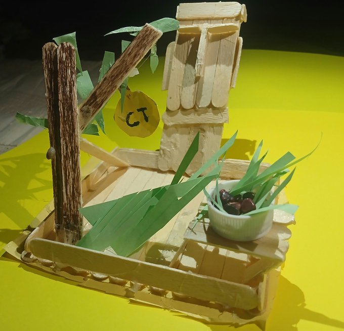 A miniature craft scene made from popsicle sticks and green paper leaves depicting a small garden with a tree and a pot of red berries on a yellow background.