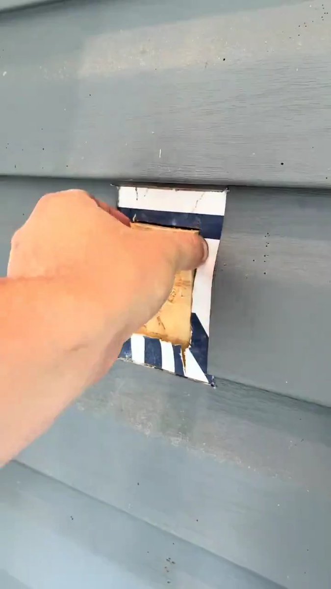 A hand rips blue painter's tape off the exposed wood framing of a freshly cut opening in gray vinyl siding, revealing meticulous diagonal masking work. The over-the-top precision of the tape job — alternating navy and white stripes protecting the surrounding siding during what appears to be a window or vent cut-in — matches the post's playful jab at the unnecessary level of care taken for a residential project.
