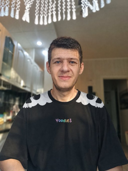 A selfie of a young man wearing a black t-shirt with "Yoodles" text and white scalloped shoulder details, taken indoors under a chandelier.