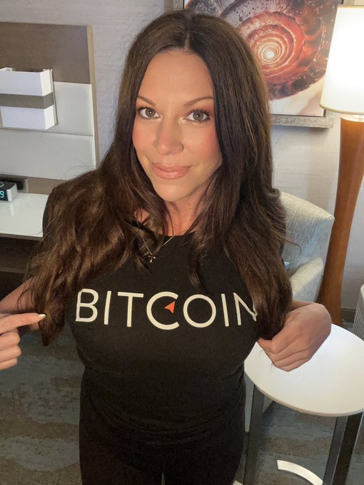 A woman with long brown hair smiles while pointing at her black "BITCOIN" t-shirt, with the "C" stylized as the Bitcoin logo, in what appears to be a hotel room setting. Her enthusiastic display of the Bitcoin-branded shirt perfectly complements her "Gm if you love Bitcoin at any price" morning greeting, showcasing her dedication to the cryptocurrency.