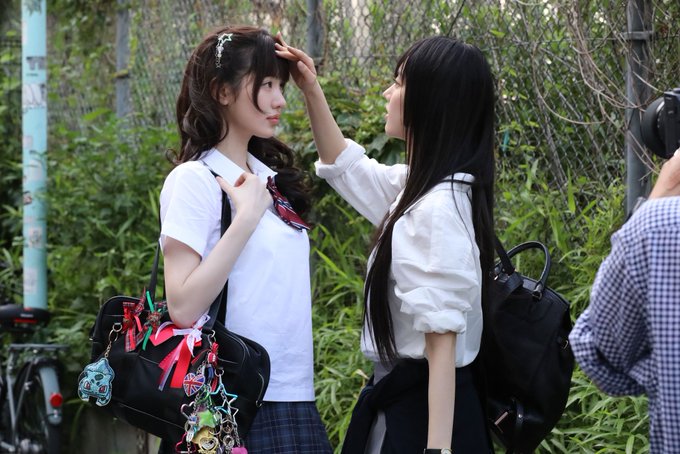 Two young women in white tops and black skirts appear to be in a cosplay photo shoot, with one adjusting the other's hair in an outdoor setting surrounded by greenery, while a photographer with a checkered shirt holds a camera on the right. The scene, tagged as "Kiki Berries #Sui" with reference number 260818, captures a tender moment between the two cosplayers.