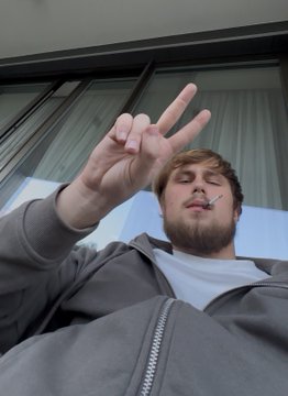 A bearded young man makes a peace sign while posing for a low-angle selfie outdoors.