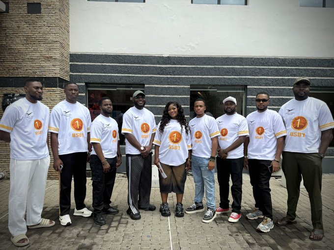 A group of nine people, including one woman, pose together outdoors wearing matching white t-shirts featuring a "1" logo and the text "usdt." The shirts reference Tether (USDT), a major stablecoin, visually connecting to the post's message about Africans using stablecoins like USDT as inflation hedges, for dollar access, and to enable faster cross-border payments.