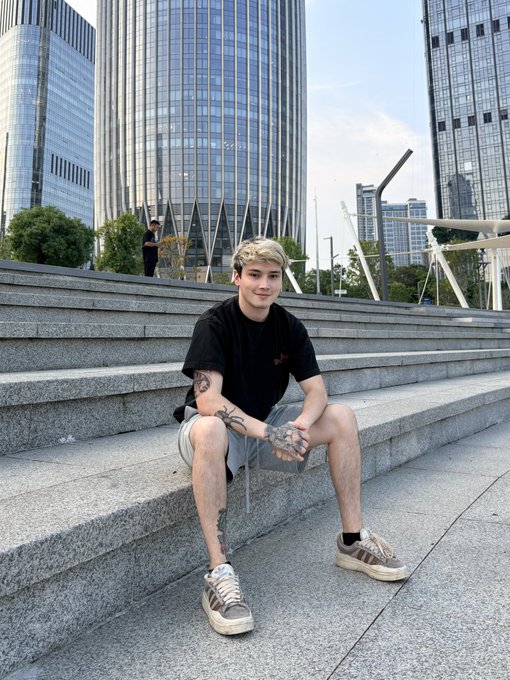 A young man with blonde hair and arm tattoos sits on stone steps in an urban plaza, with modern glass skyscrapers visible in the background.