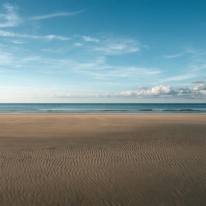 A bright, sunny beach scene featuring rippled sand patterns in the foreground, calm ocean waters, and a blue sky with scattered clouds.