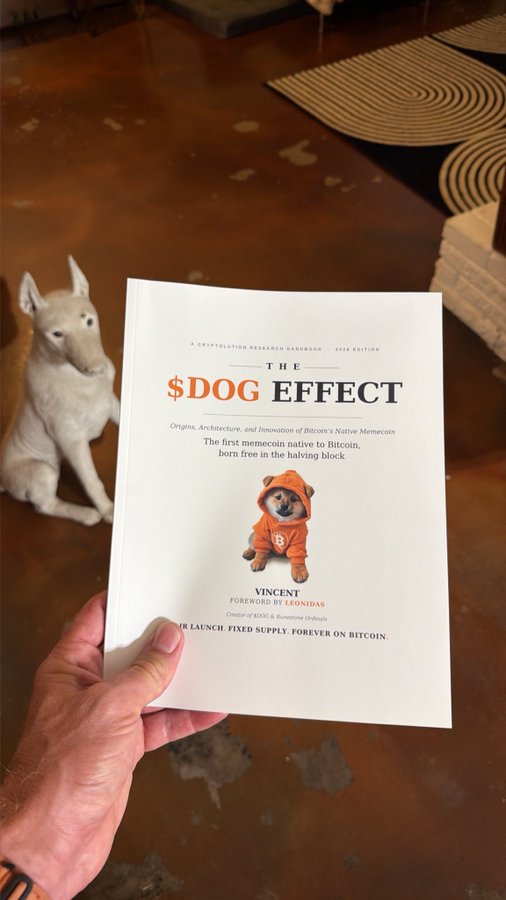 A hand holds a book titled 'THE $DOG EFFECT: Origins, Architecture, and Innovation of Bitcoin's Native Memecoin' by Vincent, with a foreword by Leonidas, featuring a Shiba Inu in an orange hoodie on the cover. A white Bull Terrier statue sits in the background on a brown floor.