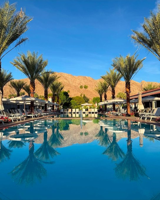 A serene resort pool scene featuring a large swimming pool reflecting palm trees and surrounding lounge chairs, with desert mountains visible in the background under a clear blue sky.