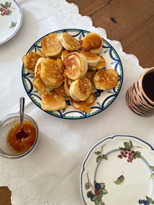 A stack of golden oladushki (small Russian-style pancakes) is arranged on a decorative blue and green patterned plate, accompanied by a bowl of homemade mango and ginger jam and a cup of hot tea, with two empty floral plates set nearby on a lace-trimmed linen tablecloth.