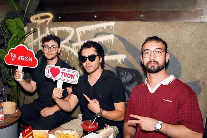 Three friends pose together at a TRON-branded event, holding "I ❤ TRON" signs in front of a graffiti-covered wall, with one person pointing to the sign and another making a peace sign, capturing the social atmosphere of the TRON TGF VIP gathering mentioned in the post.