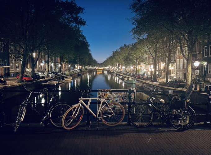 Bicycles parked on a bridge overlooking a serene canal in Amsterdam at twilight, with a sticker-covered post reading "LITE" visible among them. The European cityscape setting reflects the global and forward-looking spirit of the Litecoin Summit, embodying the optimistic message of building toward a bright future.