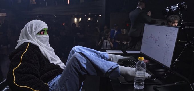 A young person wearing a white hijab and black jacket sits with their feet on a desk, looking at a computer monitor displaying stock charts, with a water bottle nearby in what appears to be a broadcast or trading environment.