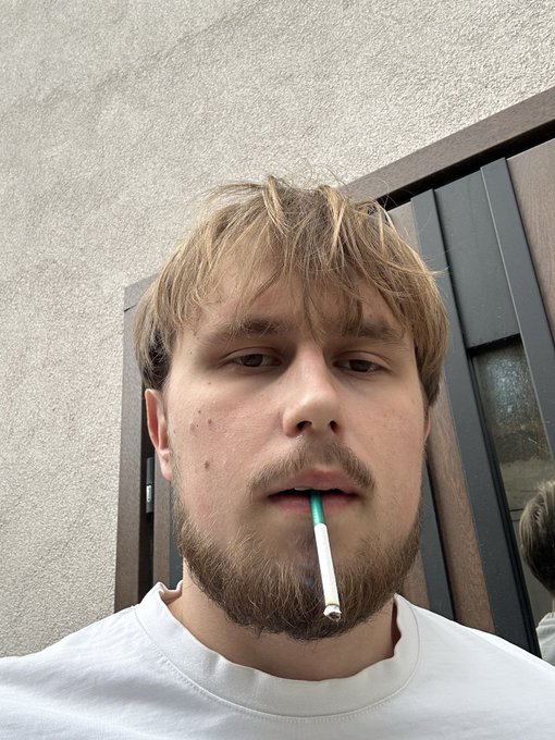 The image features a person, presumably a man or a boy, wearing a white shirt. The man has a beard and a mustache, giving him an edgy appearance. He's holding up a cigarette with his thumb and index finger, which is located in the top right corner of the photo. 

The caption to this image provides context about the day - it reads "Sunday to-do list: ..." The man appears to be engaged in some sort of activity related to social media or online content consumption, as suggested by the caption.