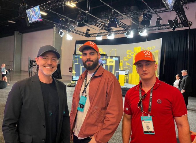 Three men pose together at an indoor event space; the man on the right wears a red polo and an orange cap with a 'Liontec' logo.