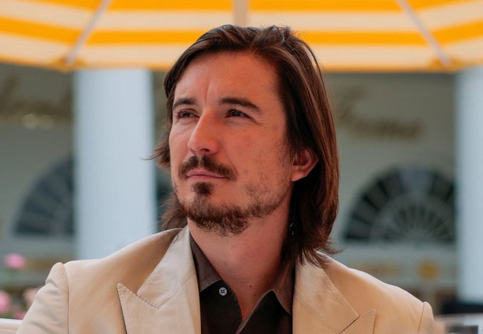A man with shoulder-length brown hair, a goatee, and a beige blazer looks off into the distance under a yellow-striped awning. The image has no connection to the post about Robinhood's crypto perps launch, as the person pictured cannot be identified by their facial features.