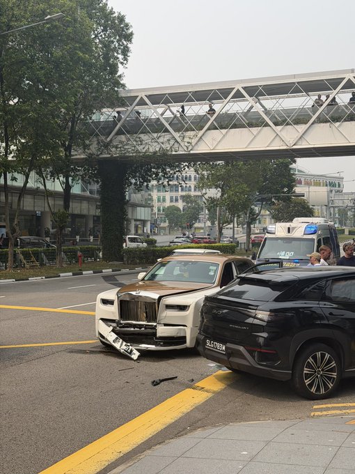 A damaged Rolls-Royce with a detached front bumper after a collision with a black Range Rover on a city street, with a license plate "SLP7634N" visible—fitting the post's description of a wrecked rental following the Token2049 event.